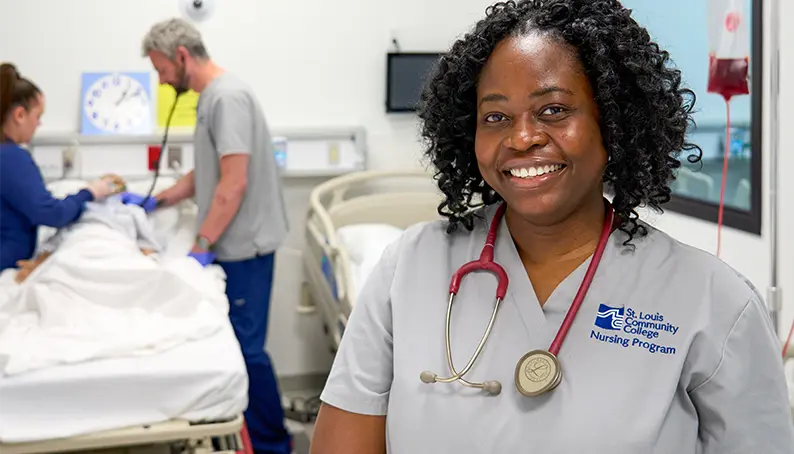 A medical student smiling for the camera with other students working on a training dummy behind her